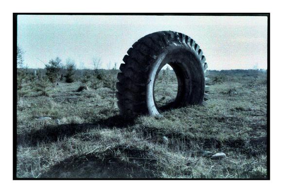 An expired film photograph of a large, weathered industrial tire standing upright in an open, grassy field. The landscape is sparse with small pine trees scattered across the terrain, under a pale, washed-out sky. The image has a grainy, bluish tint typical of old or expired color film.