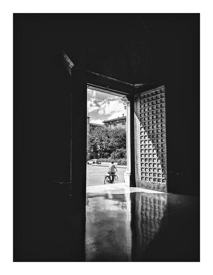 A black and white photograph of a church doorway. In the middle of the photo you can see outside the door onto a square. In the distance there are buildings, some trees and a sky with clouds. At the very entrance of the doorway there is a cyclist cycling away towards the right of the picture. The sunlight coming in is casting shadows and reflections of the wooden door and the shiny marble floor.