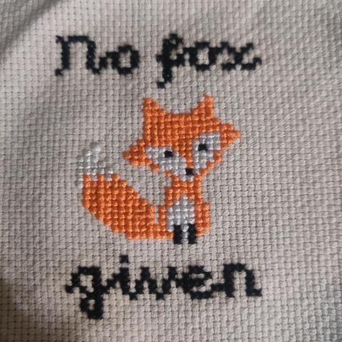 Off-white monk's cloth, cross stitched with an orange and white cartoon fox with some black in the middle. Above the fox is the text "No fox" in a cursive cross stitch script. Under the fox the text continues with the word "given".