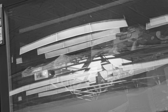 This black-and-white photograph captures a layered reflection in a glass surface, possibly a store window. Rectangular ceiling lights and structural beams from inside are overlaid with shelves of brightly lit packaged goods, visible through the reflections. The composition forms a dynamic grid of angles, lines, and transparency, creating a sense of disorientation and visual complexity.