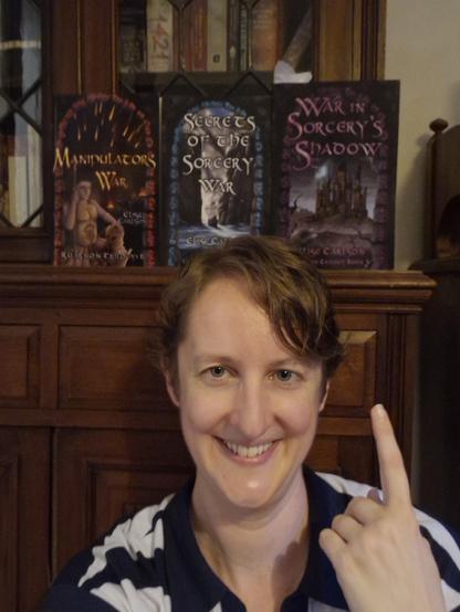 Selfie of Elise with paler than usual white as skin because I'm sick, slight pink in cheeks and massive smile, my index finger pointing up the three books in my Ruarnon trilogy, lined across the shelf above the cupboard section, and before the lead light glass level of my antique bookshelf. Proper covers alt text in next image.