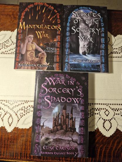 Ruarnon Trilogy books arranged; Manipulator's War, Secrets of the Sorcery War above, War in Sorcery's Shadow below on large lacy table runner and ancient grained antique side table.

Manipulator's War Cover
Left: portrait of young Heir Ruarnon, their hair braided back, eyes outlined with kohl, wearing fitted bronze armour, a bronze disc kilt, holding a helmet in their left hand and leaning on a spear with their right, which pierces the ‘A’ in the title.
Right: Red glyphs outline a stone archway, through which fire arrows rain down on torch-lit battlements atop a castle, at night time. 
Bottom text: Elise Carlson, Ruarnon Trilogy Book 1
 
Secrets of the Sorcery War
Blue glyphs in a stone arch frame a pair of tall cliffs, a ship sailing on bright blue water through the gap, into the sunshine of a natural harbour.
Bottom text: Elise Carlson, Ruarnon Trilogy Book 2
 
War in Sorcery's Shadow
Purple glyphs around a stone arch in a stone wall, through which blackened clouds, with grey, lightening pronged edges loom over a many brown towers castle. Lightning crashes against and partially reveals a magic dome shield around the castle.
Desolate hills and dray plains life before it, with leafless, dried-up trees and a skull in the foreground.
Bottom text: Elise Carlson, Ruarnon Trilogy Book 3