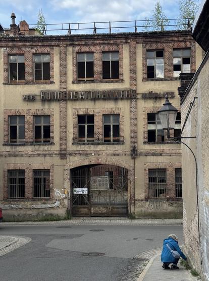 Ein verlassenes Fabrik-Gebäude mit dem Schild „VEB Kondensatorenwerk Görlitz“ an der Fassade. Die Struktur verfügt über mehrere Fenster, von denen einige kaputt sind, und einen geschlossenen Eingang. Eine Person in einer blauen Jacke hockt in der Nähe des Bürgersteigs und zupft wilde Pflanzen aus dem Boden