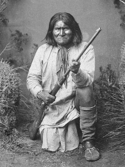 Photographie en noir et blanc de Geronimo, chef de guerre apache Chiricahua, prise en 1887. Il est agenouillé sur un sol herbeux, tenant un fusil dans ses mains. Il porte une tunique traditionnelle avec des mocassins et des jambières. La scène est entourée de végétation, avec des plantes et des buissons visibles en arrière-plan. Cette image historique illustre la résistance des Apaches face aux forces américaines et mexicaines.