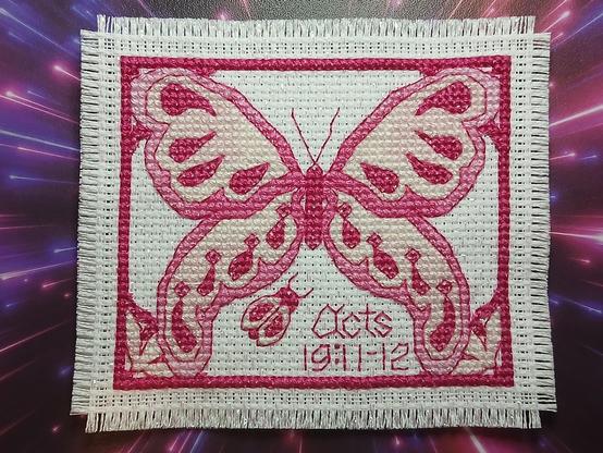 This is a picture of the front panel of "Blue Willow Butterfly Prayer Cloth in Pink." It shows a cross stitched butterfly with a border, two flowers in the lower corners and a #Ladybug, all in pinks. The ladybug is positioned just to the left of the center toward the bottom below the butterfly's lower left wing. To the right of the ladybug, "Acts 19:11-12" is stitched, also in pink. The background for the photograph is actually my mouse pad. It has what looks like rays of pink and purple light, making it appear that they're streaming toward the viewer as if the light is coming from behind the prayer cloth.
