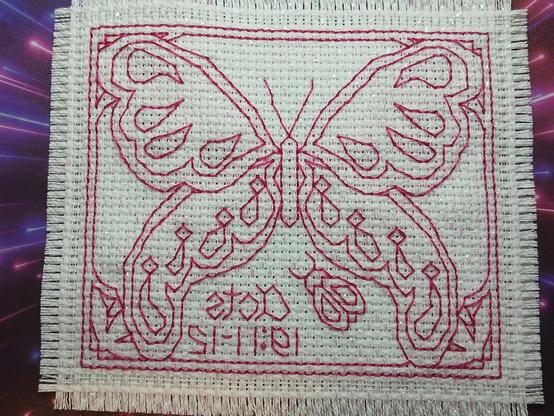 This is the back panel of "Blue Willow Butterfly Prayer Cloth in Pink." The picture shows the outlines of a butterfly with a border, two flowers in the lower corners and a ladybug, all in a dark  pink, almost as if it is a line-drawn mirror image of the front panel. The ladybug is positioned just to the left of the center toward the bottom below the butterfly's lower left wing. To the right of the ladybug, "Acts 19:11-12" is stitched, also in pink. The background for the photograph is actually my mouse pad. It has what looks like rays of pink and purple light, making it appear that they're streaming toward the viewer as if the light is coming from behind the prayer cloth.