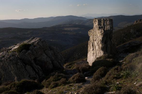 On se trouve dans un paysage grec où se succèdent une série de montagnes arides. Sur le haut de l’image, à l’horizon, une bande de ciel bleu clair. Au premier plan, sur la droite, une roche dressée, aux angles presque droits, reflète la lumière matinale du soleil, et contraste avec le reste du paysage.