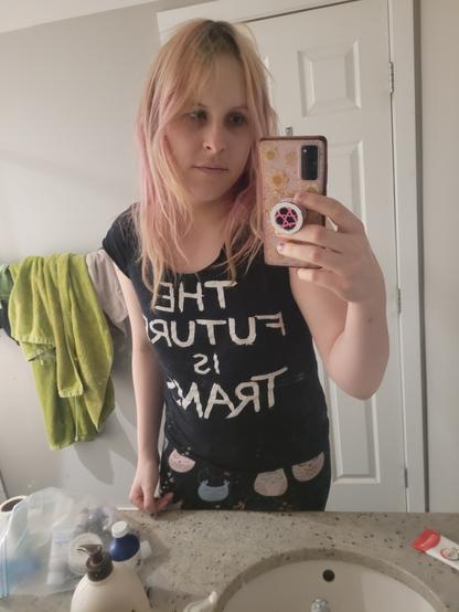 tired trans girl taking a bathroom mirror selfie. she has pink hair that's fading into bleach blonde at the roots and a shirt that says, the future is trans