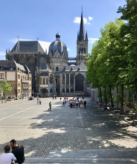 Blick vom Rathaus aus über den Katschhof auf den Aachener Dom. Am rechten Bildrand sind grüne Bäume zu sehen, auf dem gepflasterten Platz ist eine Touristengruppe zu erkennen. Vorne links hat sich ein Pärchen auf Stufen niedergelassen. Der Himmel ist blau und sommerlich.