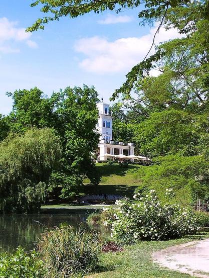 A white tower peeks through lush green trees, standing beside a serene pond. The surrounding gardens are vibrant with various plants and bushes under a clear blue sky.