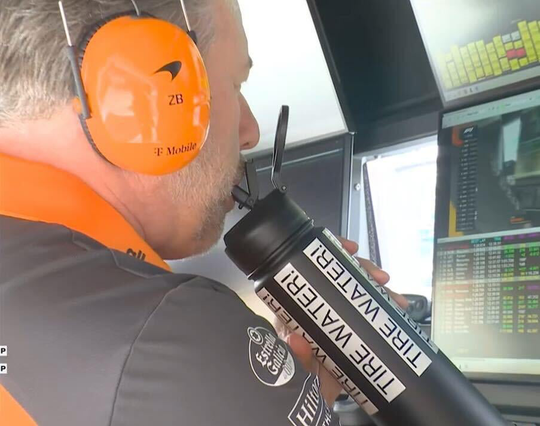 Zak Brown watching a screen and drinking from a bottle with multiple "Tire Water!" stickers on it.