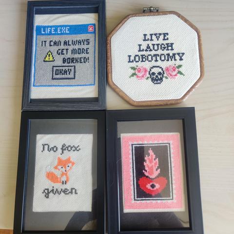Four finished cross stitch pieces, three of them framed in photo frames behind glass and one of them framed in a fake wooden octagonal hoop-frame.

One is a flaming heart with borders, colours used: red, black, white, bright pink, dark pink.
One is a fox with the text "no fox given", colours used are orange, white and black.

One is a windows xp style error box saying "warning, it can always get more borked" with a caution sigh next to the text and an "okay" button under the text.

The one in the special frame says "live laugh lobotomy" with a skull and mirrored flowers at the bottom.