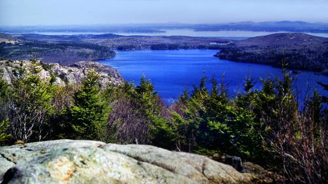 Beneath a clear blue sky we are standing on a light gray granite outcrop atop a mountain, looking out over the tops of a mixed forest of evergreens and leafless hardwoods running down the mountainside before us. Just beyond the trees on our left and just below us is a rocky crag with only a few stunted pines growing from crevices along its crest. A large deep blue pond dominates the view below in front of us. To our right a small rounded mountain rises from the pond's shore. Beyond the pond is a narrow, flat wooded strip of land backed by a long ocean inlet and then another relatively fat piece of land  with a range of mountains to our right and scattered mountains along the horizon.