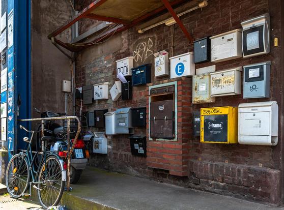 Eine Wand mit vielen völlig verschiedenen Briefkästen, die auch in völlig durcheinander aufgegangen sind. 
Davor stehen ein Motorroller und ein Fahrrad.