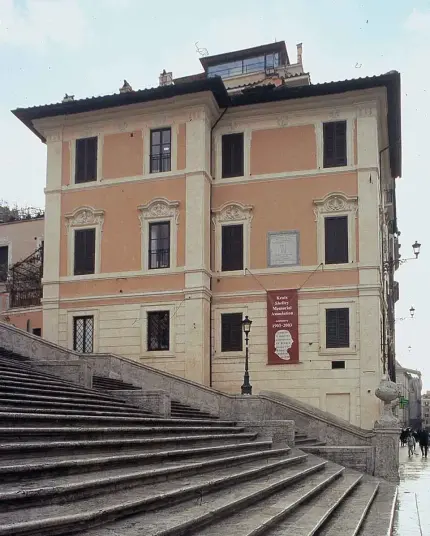 The image features a building with a distinct architectural style, likely from the historical period. It appears to have multiple windows and is designed with decorative elements. A banner is displayed on the building, and there are steps leading up to it, suggesting it is situated in a public area, possibly near a famous landmark or square. The surrounding environment looks urban and is likely to be in a city known for its history and architecture.

Image Credits: Wikimedia / Keats1795 / Public domain