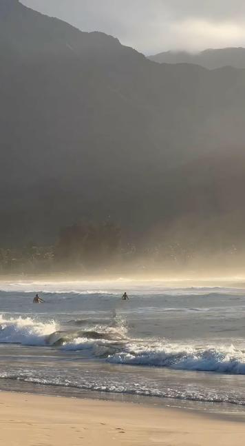 A serene morning scene with surfers, out in the waves, gliding over calm waves, the view is framed by the soft light of dawn at the ocean's edge. Stunning cloud covered green mountains beyond the shoreline. The view capturing the essence of relaxing time at the ocean.