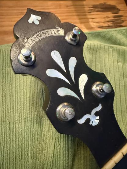 The head of a banjo without any strings through the tuning pegs, showing the newly cleaned teardrop inlay patterns and metal makers mark that looks like a ribbon and reads LANGSTILE.