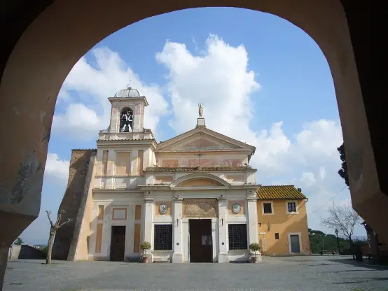 The image shows a building that appears to be a church or a historic structure, framed by an archway. The architecture features a bell tower and some decorative elements on the façade. The sky is blue with scattered clouds, and there are a few trees and a yellow building in the background. The setting looks to be outdoors, with cobblestone pavement visible in the foreground.

Image Credits: Wikimedia / Lalupa / CC BY-SA 3.0