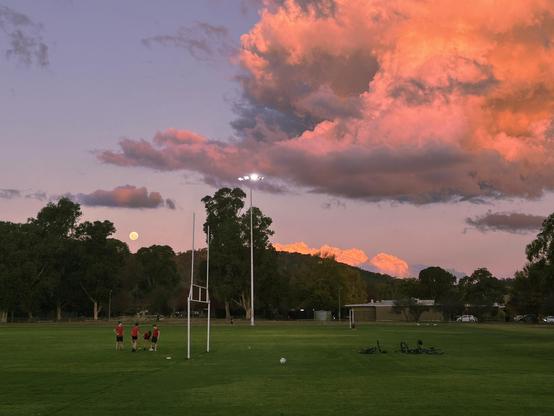 Photo of a school oval with kids playing underneath a full moon just above some trees with a large, brightly coloured cloud in the sky.