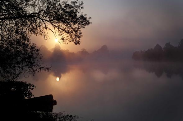 Le soleil se lève au dessus d’une rivière enveloppée dans la brume. La lumière est rouge orangée. Sur la gauche, la branche d’un arbre sous laquelle pointe le soleil, et plus bas, la silhouette de l’extrémité d’une barque. Un peu au-dessus de la moitié de l’image, la traversant, on devine les arbres qui bordent le cours d’eau. On dirait presque un petit lac.