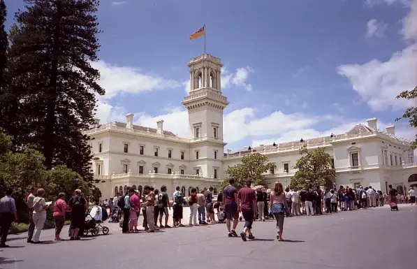 The image shows a large, grand building, likely a government or historical structure, characterized by its white exterior and prominent tower. There is a flag flying from the tower. In front of the building, there is a long line of people waiting, likely to enter or visit the location. The setting appears to be a sunny day with some lush greenery around.

Image Credits: Wikimedia / Photograph by Greg O'Beirne / CC BY 2.5