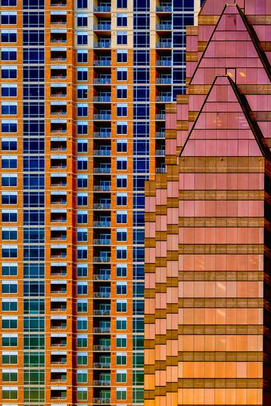 A vertical image of two buildings. On the left, a tall building stretches upwards, its facade a grid of warm, reddish-brown panels punctuated by deep blue rectangular windows. Each window reflects the sky, creating a rhythmic pattern. Thin, light-colored horizontal lines divide the floors. Some floors feature small balconies. To the right, a second building rises, its surface a shimmering blend of pink and copper hues. This building has a more angular, almost stepped design, with strong diagonal lines converging towards the top. The light reflects off its glass panels, creating a sense of depth and movement. The two buildings create a contrast between the sharp angles of the right buidling with the more uniform grid of the left building.