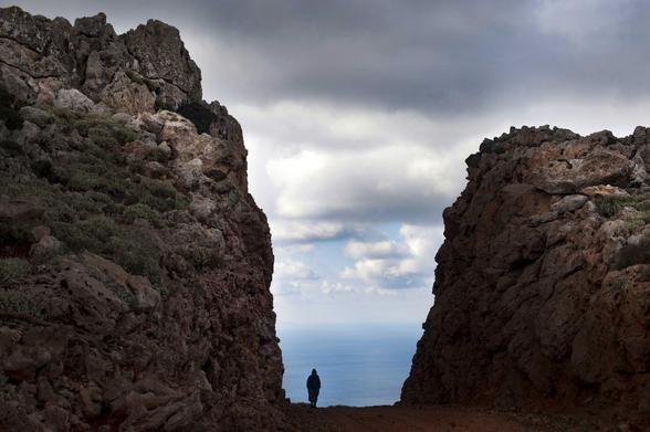 Un chemin de terre coupe en deux une sorte de colline rocheuse qui descend en pente douce de la gauche vers la droite. Une personne se trouve sur le chemin, juste entre les deux pans de rochers: c’est une petite silhouette qui se détache de la mer qu’on voit au-delà du chemin, et qui rejoint le ciel au premier tiers de l’image pour former l’horizon. Le ciel est nuageux.