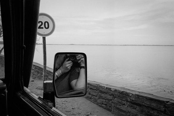 a black and white photo of a person taking a photo out of car on the passenger-side, the person and camera are visible in the side-mirror
