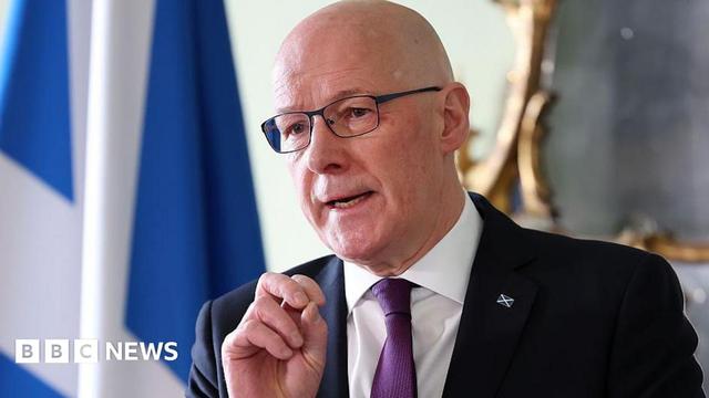 John Swinney, a bald man with glasses wearing a dark suit and purple tie, with a saltire pin on the lapel. He is standing in front of a saltire flag during a press conference mid speech with his hand elevated.