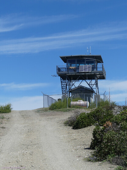 Just one level worth of steps leads up to the observation deck of a fire lookout with the shutters open. A sign identifying it as Horse Ridge Lookout and solar panels decorate the railing. There is a shed and propane tank underneath and a chain link fence surrounds it. A dirt road climbs up to the peak beside it on the left. All is under a sky of long wispy clouds across the blue.