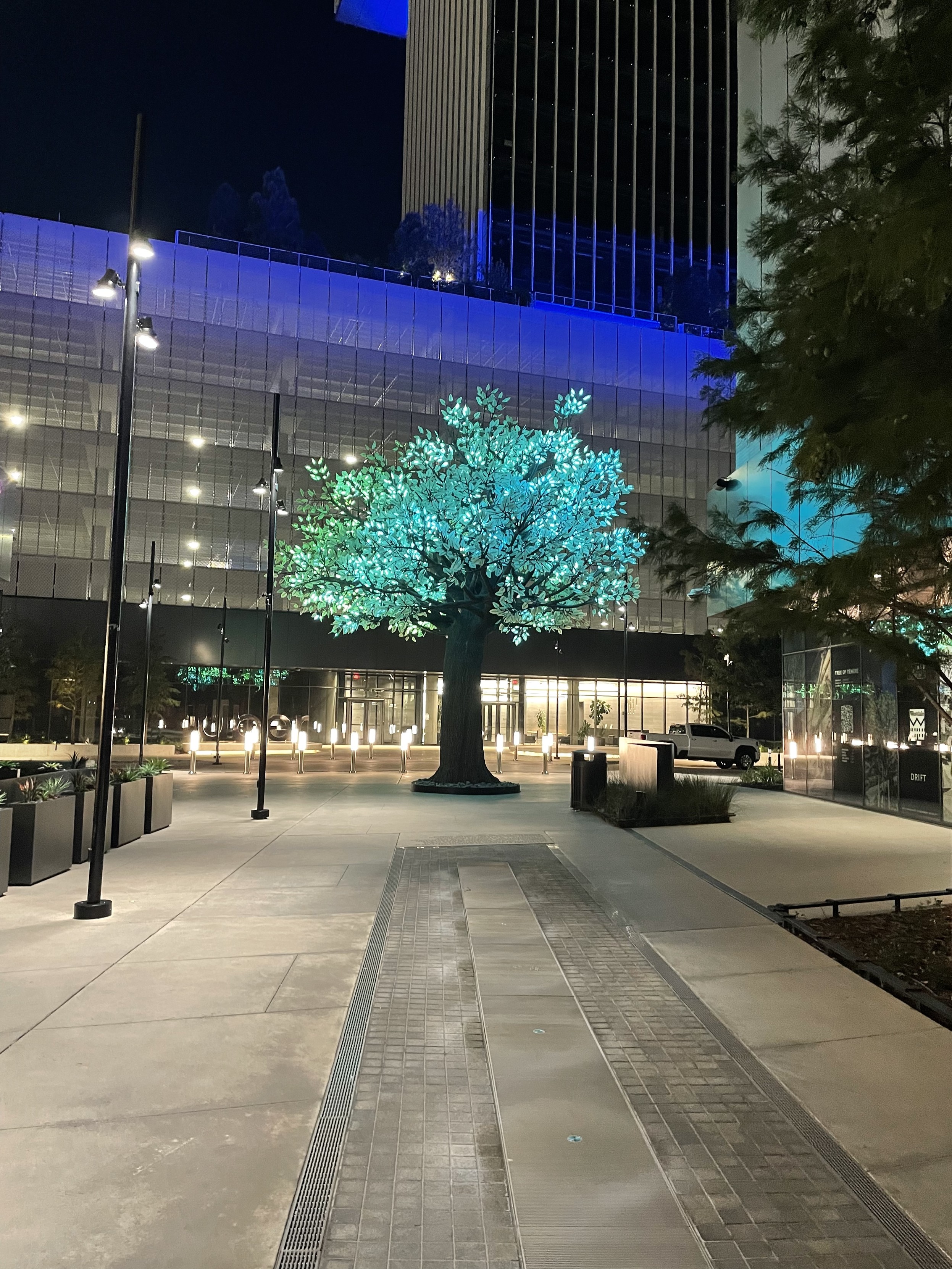 An artificial tree made out of steel and aluminum. It has a very thick canopy made of leaves made up of LED lights. 
In this picture, the lights are currently an aqua blue color. 

Behind the tree is a parking garage and a high rise building. 

The night sky is seen in the background. 