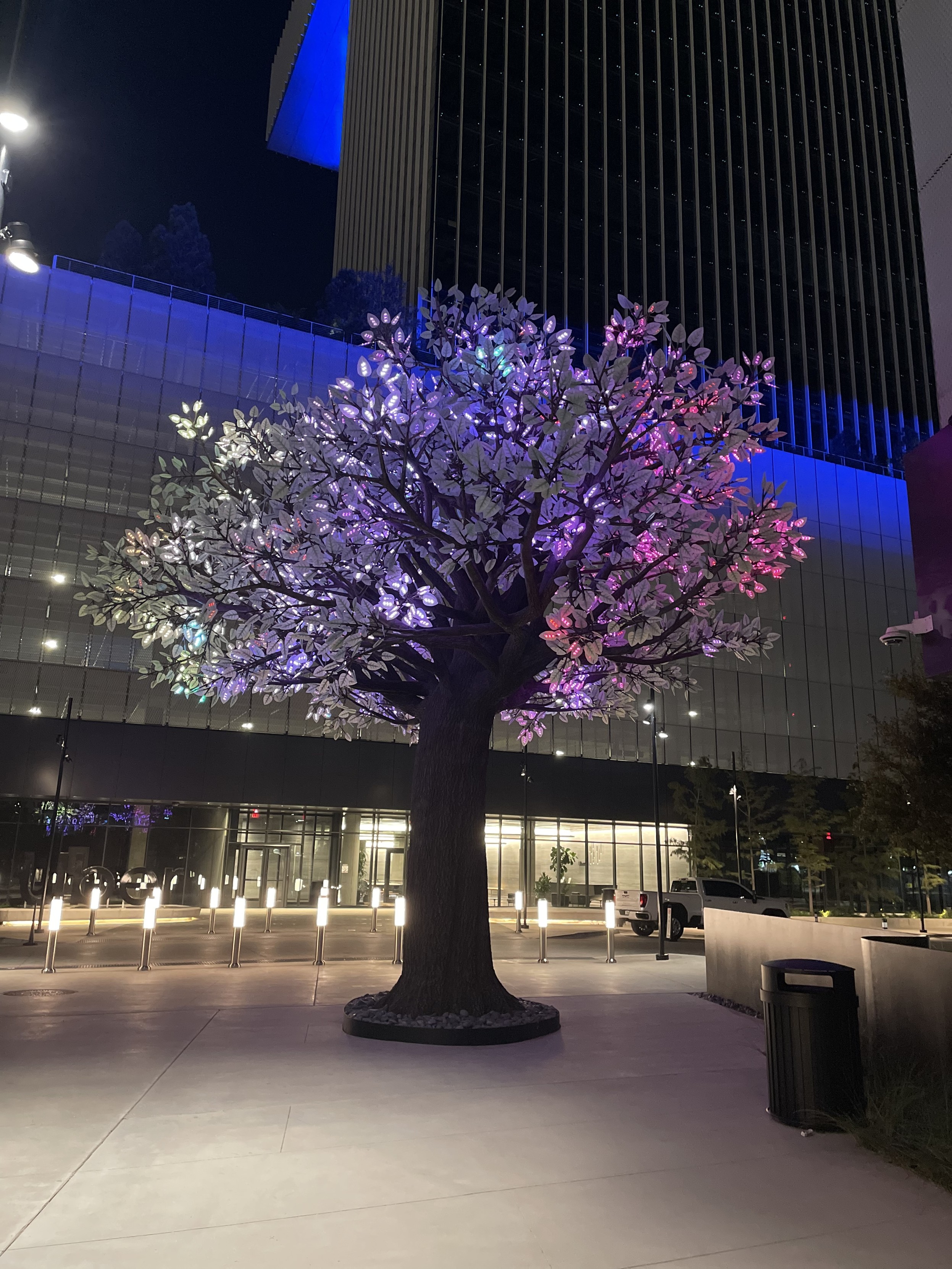 An artificial tree made out of steel and aluminum. It has a very thick canopy made of leaves made up of LED lights. 
In this picture, the lights are currently white and various shades of purple. 

Behind the tree is a parking garage and a high rise building. 

The night sky is seen in the background. 