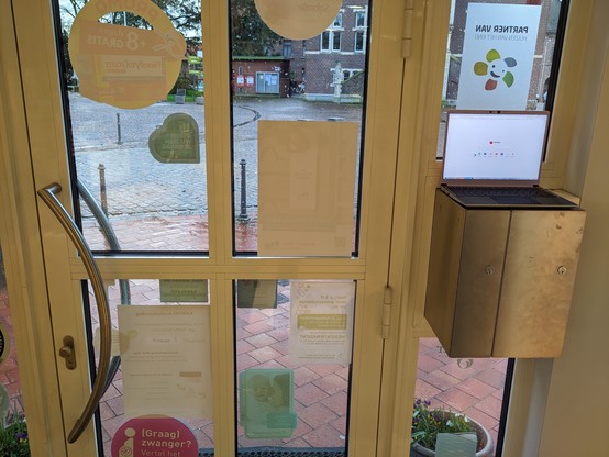 The image shows the front door of a pharmacy from the inside looking out. The glass door has multiple flyers and notices attached.
Through the door's windows, a cobblestone street and a few parked bicycles are visible, indicating a quiet outdoor scene. To the right inside, a bronze-colored metal mailbox is mounted on a cabinet, above which a laptop is open on Firefox
