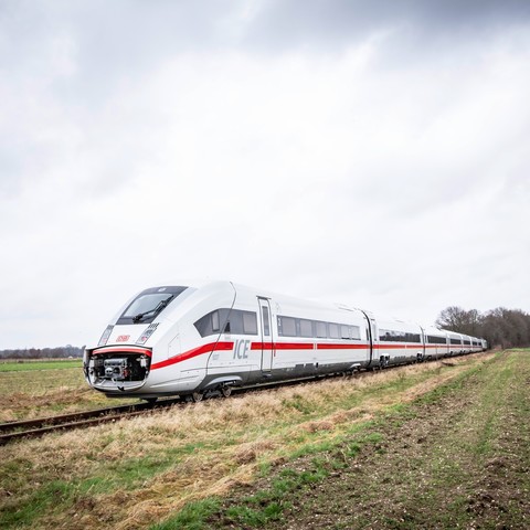 A sleek white and silver high-speed train with red accents and the "ICE" logo, stands on open tracks amidst a rural landscape under a cloudy sky.