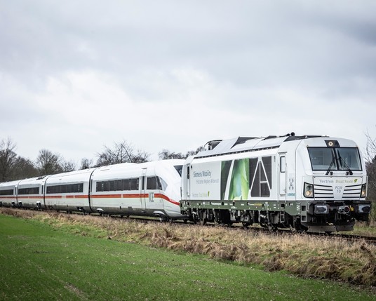 A modern dual-mode locomotive in white with green and black graphics, labeled "Siemens Mobility Vectron Dual Mode," is coupled to the high-speed train carriages in a natural setting with leafless trees in the background.