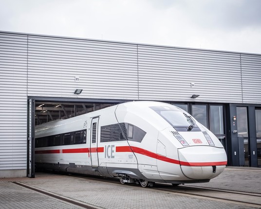 The front carriage of a high-speed ICE train, predominantly white with red and silver accents, is partially inside a maintenance hangar with a corrugated metal exterior. Numbers "84" and "85" are visible on the hangar wall above the train.
