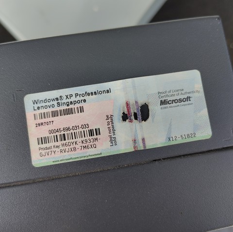 A worn label on a device displays a Windows XP Professional license sticker. It indicates Lenovo Singapore as the manufacturer with a partially scratched product key. Visible characters in the key are "H6DYK-K933M-GJV7Y-RJVX8-7M6XQ". The sticker includes Microsoft's holographic security features and a URL for anti-piracy information, with text "Proof of License Certificate of Authenticity" and "©2005 Microsoft Corporation". There's slight damage where parts of the product key are scratched out.

