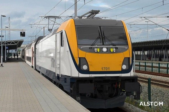 A modern TRAXX 3 Multi-System (MS) locomotive numbered 1701 is stationed at a train platform. The locomotive features a sleek design with a yellow and gray front and a white body. The platform is paved with light gray tiles, and there are overhead power lines and signals visible above the train. To the left, part of the station infrastructure, including a canopy and waiting passengers, is visible. The Alstom logo is present in the lower right corner of the image.

