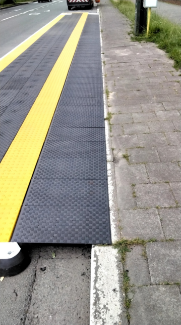 A close-up view of the temporary bus stop platform showing the tactile black and yellow paving strips running parallel to the curb. The platform is positioned next to a brick-paved sidewalk, which has some grass growing between the bricks.