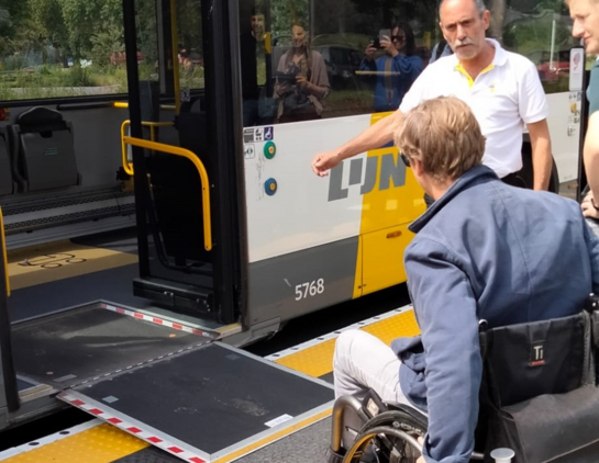 A man in a white shirt and several other individuals are assisting a person in a wheelchair to board a bus from the temporary platform. The bus has a ramp extended from the door to the platform, allowing smooth boarding. The platform features yellow and black tactile paving, and the bus is marked with the number 5768.
