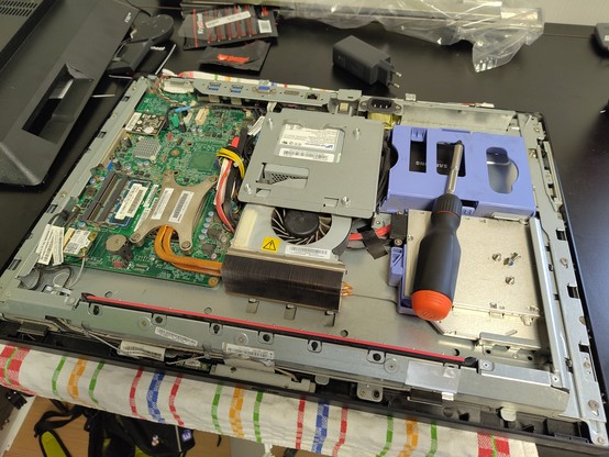 An open all-in-one PC with its internal components exposed, placed on a table. Various parts including the motherboard, cooling system, and power supply are visible. A screwdriver is resting on the PC. The table also has other tools and components scattered around.