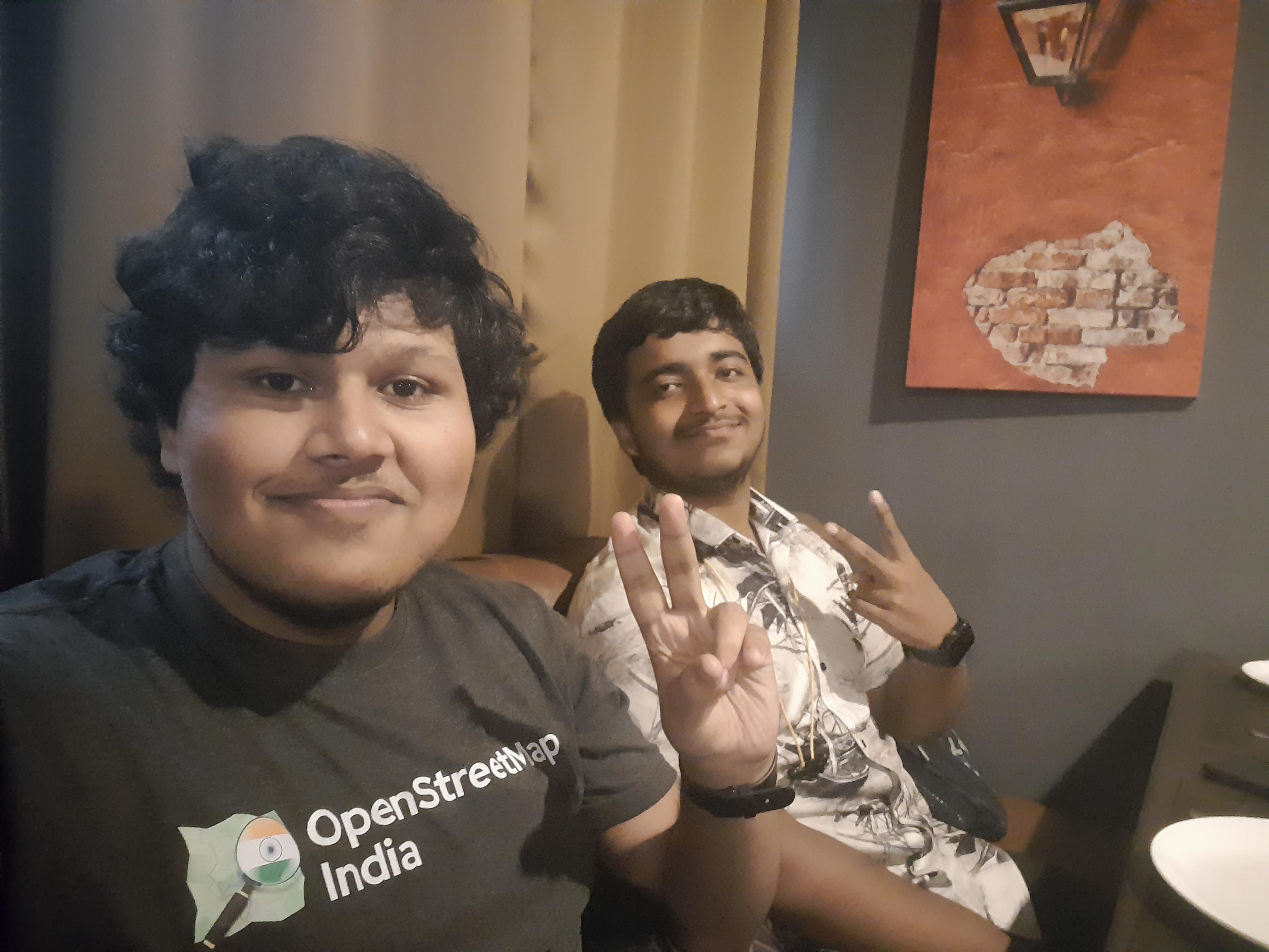 2 men sitting inside a restaurant in Siliguri, facing the camera, raising one hand doing the "V" gesture. The person in the left of the picture is wearing a grey OpenStreetMap India T-shirt. 2 men sitting inside a restaurant in Siliguri, facing the camera, raising one hand doing the "V" gesture. The person in the left of the picture is wearing a grey OpenStreetMap India T-shirt.