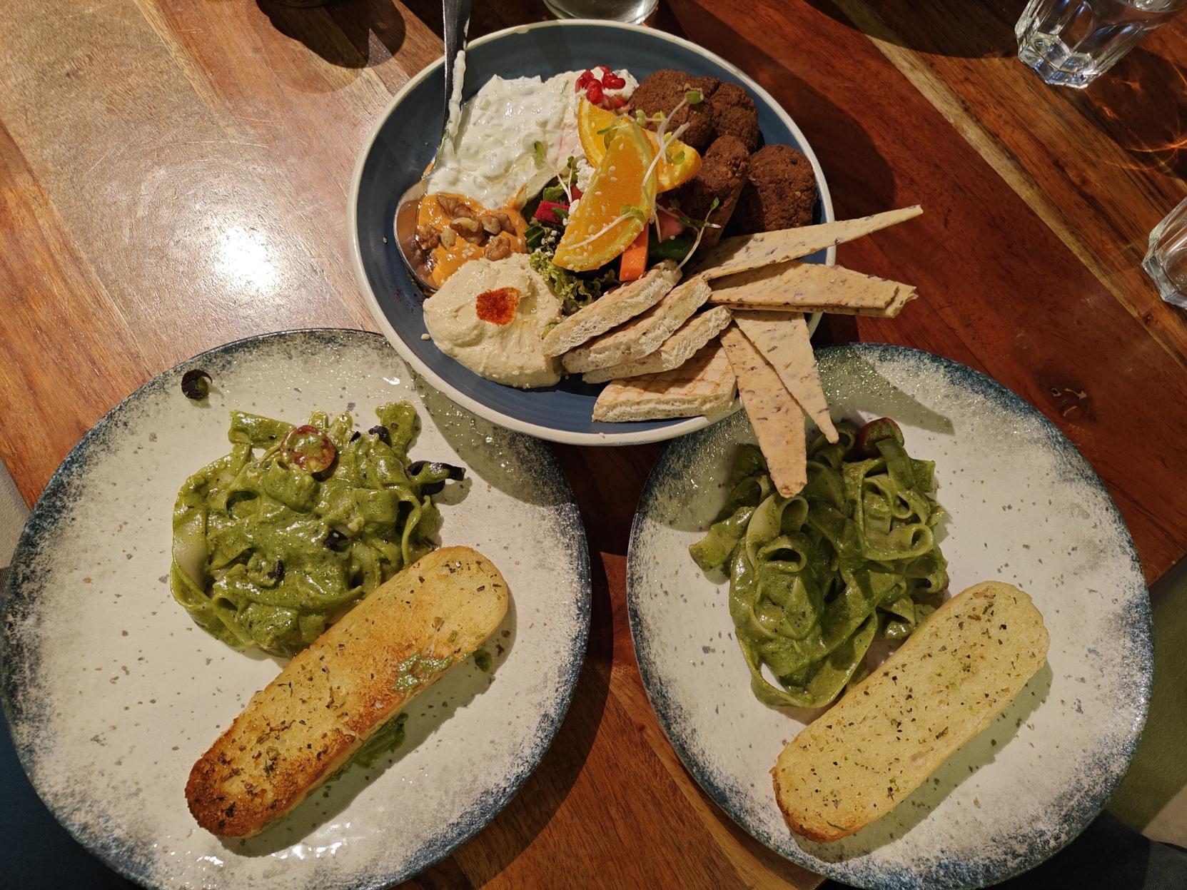 A photograph of food on a wooden table at HONS All Day Dining.
There are two plates, each with green basil pesto pasta, half a cherry tomato, and toasted oregano bread.
There's also a mezze bowl, containing toasted pita bread cut into quarters, flatbread crackers, falafel, hummus, an orange condiment, a white slaw with pomogranate seeds, another whitish condiment, slices of orange, and leaves of lettuce. A photograph of food on a wooden table at HONS All Day Dining.
There are two plates, each with green basil pesto pasta, half a cherry tomato, and toasted oregano bread.
There's also a mezze bowl, containing toasted pita bread cut into quarters, flatbread crackers, falafel, hummus, an orange condiment, a white slaw with pomogranate seeds, another whitish condiment, slices of orange, and leaves of lettuce.
