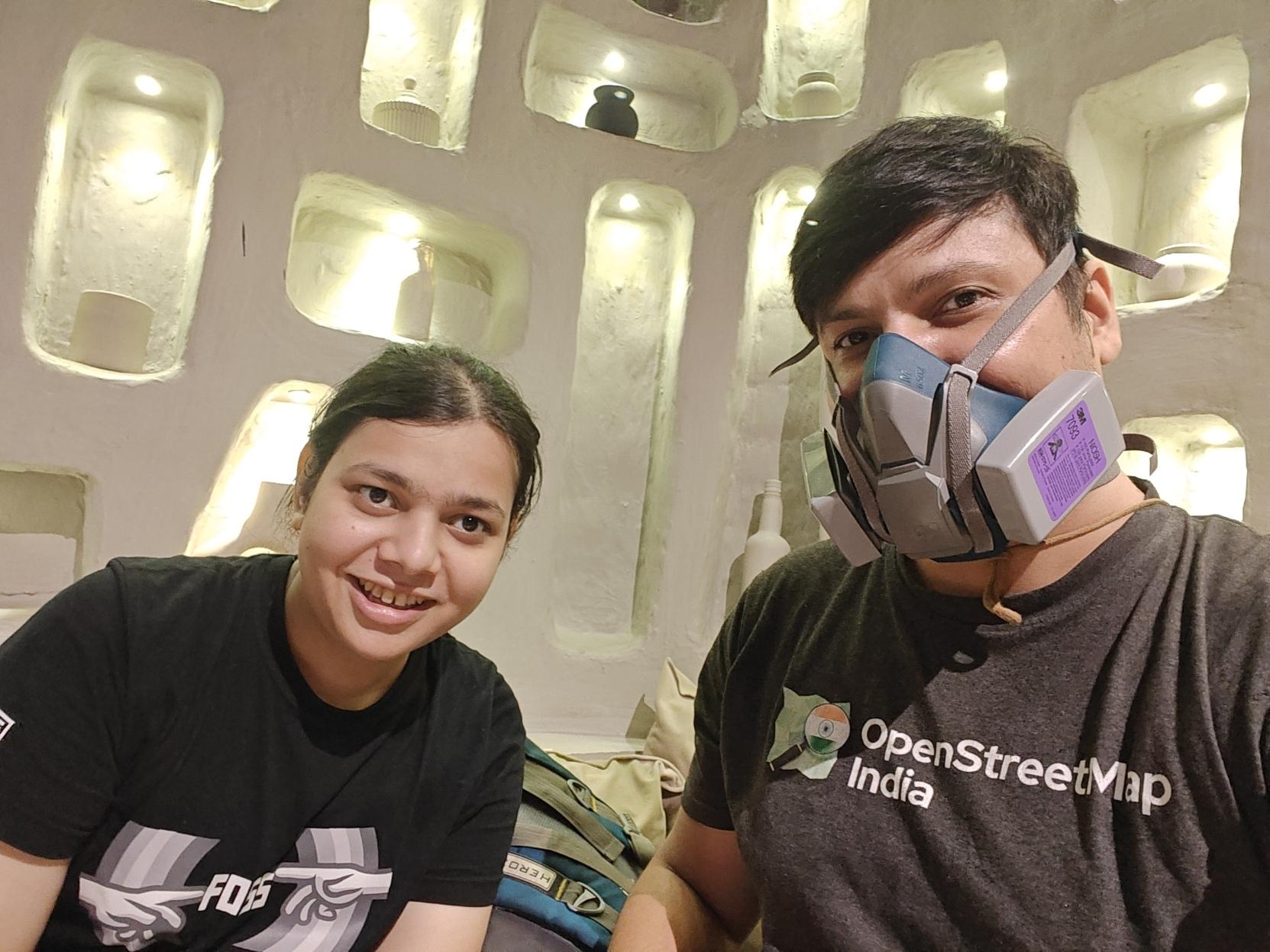 Selfie of niyabits and contrapunctus.
Niya is a fair transgender girl with long black hair tied back into a ponytail. She is wearing a black FOSS United T-shirt. The T-shirt has a graphic with two hands pointing at each other with their index fingers (similar to Michelangelo's fresco, "The Creation of Adam"), with the text "FOSS" shown between them. She is smiling at the camera.
contrapunctus is a person with messy black hair which is short at the sides and long at the top. They are wearing a grey OpenStreetMap India T-shirt, and a blue-grey 3M 6502QL elastomeric respirator with grey-and-purple 3M P100 7093 filter cartridges.
There is a plaster wall behind them with rounded-rectangular "windows" of different sizes, each containing a ceramic vessel, and a little light at the top. Selfie of niyabits and contrapunctus.
Niya is a fair transgender girl with long black hair tied back into a ponytail. She is wearing a black FOSS United T-shirt. The T-shirt has a graphic with two hands pointing at each other with their index fingers (similar to Michelangelo's fresco, "The Creation of Adam"), with the text "FOSS" shown between them. She is smiling at the camera.
contrapunctus is a person with messy black hair which is short at the sides and long at the top. They are wearing a grey OpenStreetMap India T-shirt, and a blue-grey 3M 6502QL elastomeric respirator with grey-and-purple 3M P100 7093 filter cartridges.
There is a plaster wall behind them with rounded-rectangular "windows" of different sizes, each containing a ceramic vessel, and a little light at the top.
