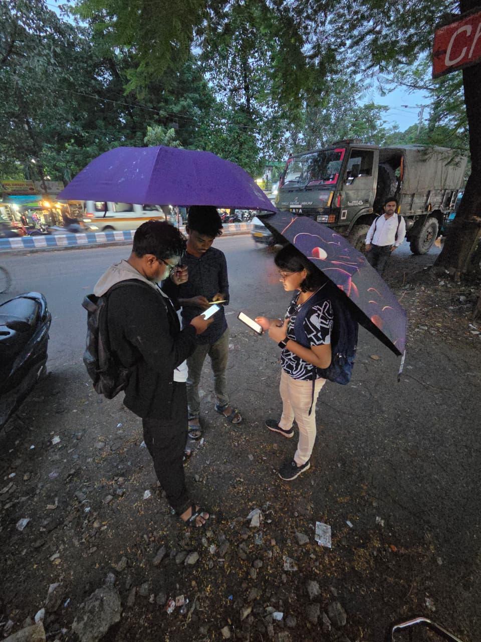 3 people, 2 men and a woman, holding umbrellas by a high street, mapping features using their smartphones.