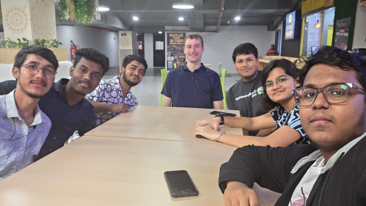 7 people, 6 men and a woman, one of the men wearing a grey OpenStreetMap India T-Shirt, sitting inside a food court at a shopping centre, smiling towards the camera.
