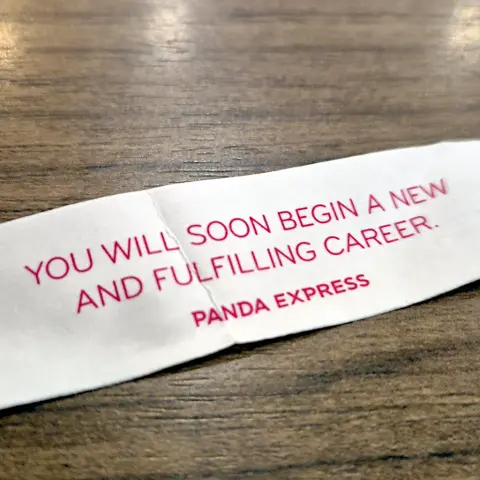 A strip of white paper on a wood-textured surface with red text stating “You will soon begin a new and fulfilling career” with “Panda Express” under it.