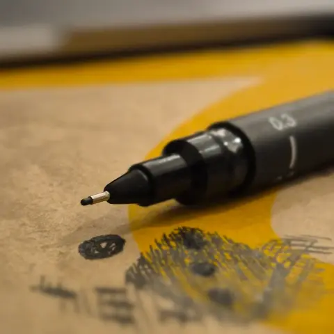 Macro photo of a gel pen in focus placed on top of brown paper with a crude drawing of a die