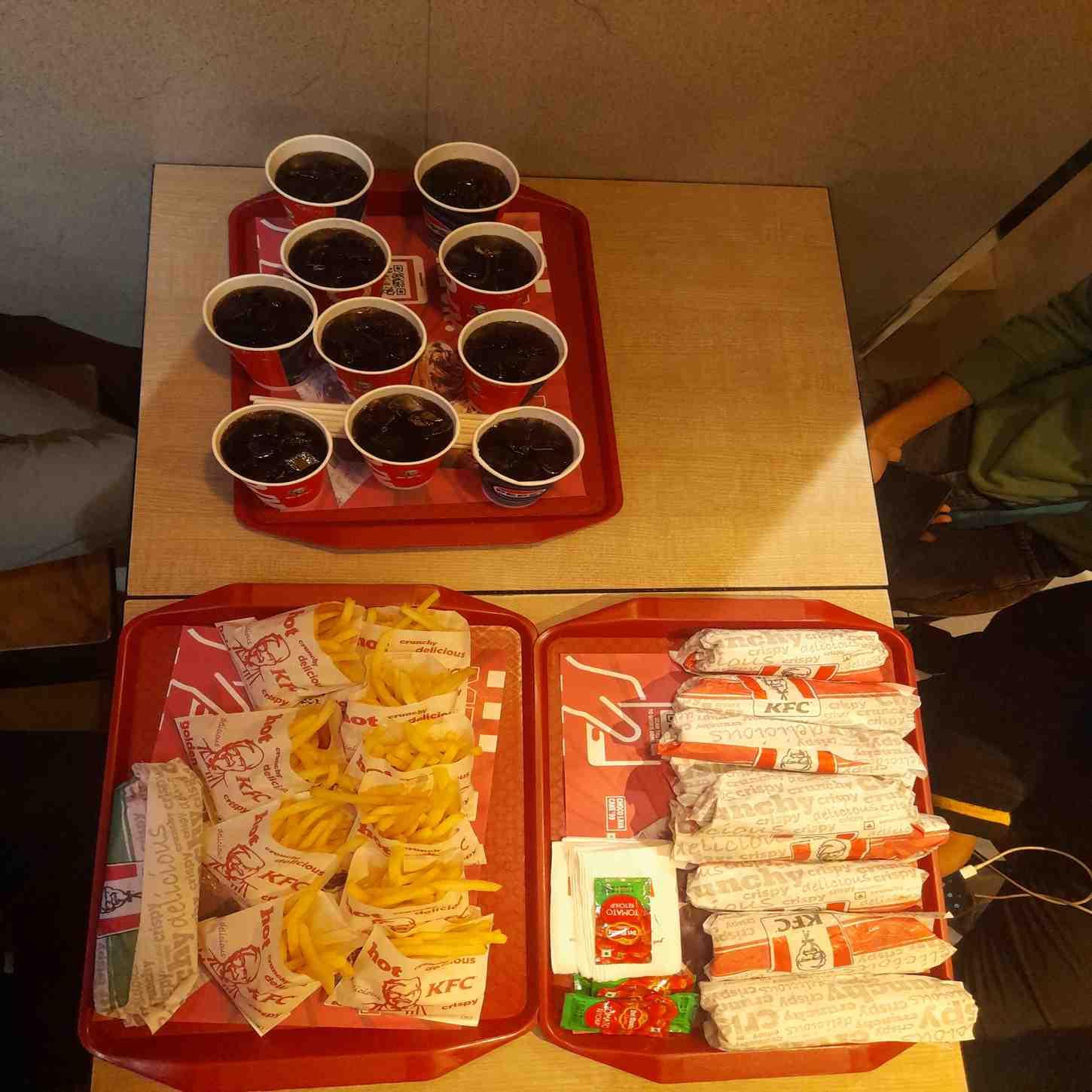 A slightly warped image of a table with 3 trays of food. One of them contains cups filled with pepsi, another with portions of french fries and a veg wrap, and the other tray with chicken wraps, and sachets of tomato ketchup and wipes.