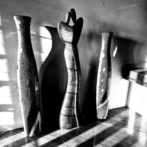 High-contrast black and white photo of three tall objects harshly lit by a horizontal light source from the left with grill shadows bathing the scene and leaving sharp lines on a wooden floor. The middle object looks like a thin abstract sculpture of two figures embracing while the other two objects on either side look like tall thin vases.